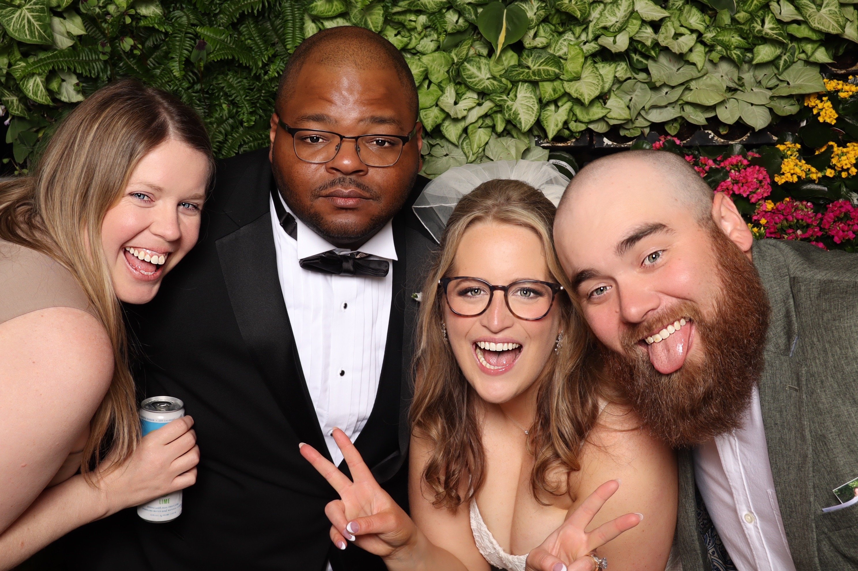 A bride and groom crowded into one frame with their guests at a greenery wall